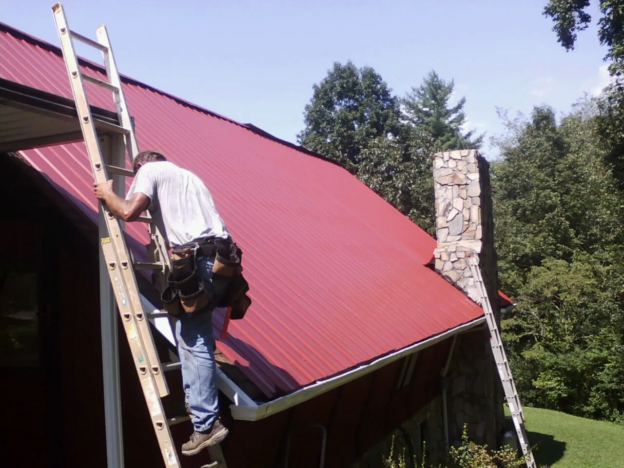 Roofer on a ladder beside a red metal roof and stone chimney — Soffit & Fascia Repair by Roofing Brothers in Mechanicstown